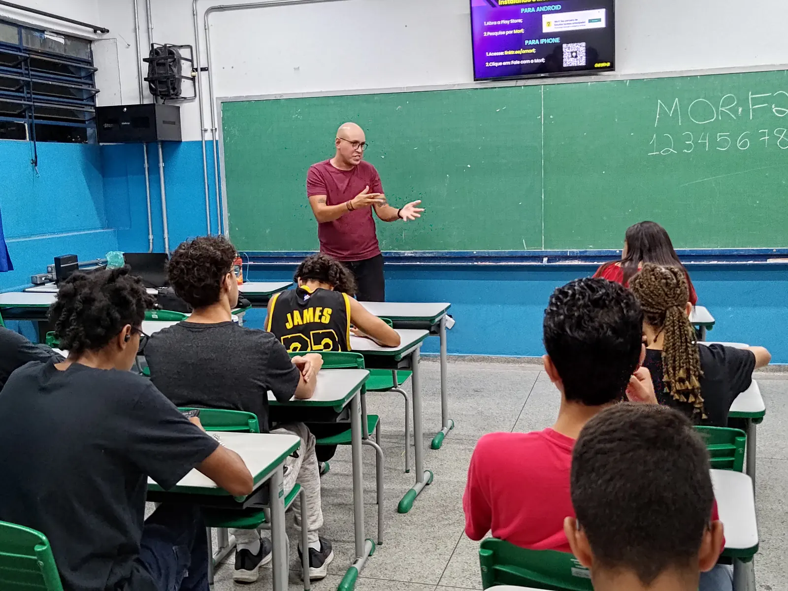 Palestrante em sala de aula com MORF escrito na lousa e slide de instalação do app na TV